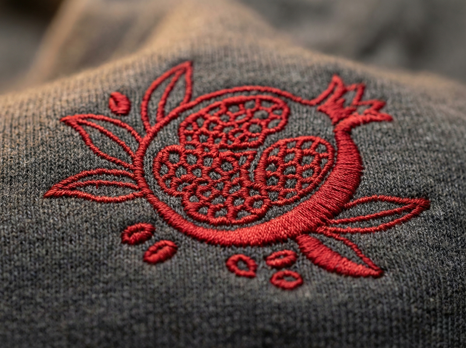 a close-up view of a gray fabric with a red embroidered design on it. The design appears to be a pomegranate or similar fruit, with intricate patterns and details.