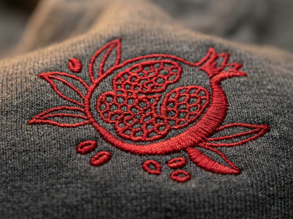 a close-up view of a gray fabric with a red embroidered design on it. The design appears to be a pomegranate or similar fruit, with intricate patterns and details.