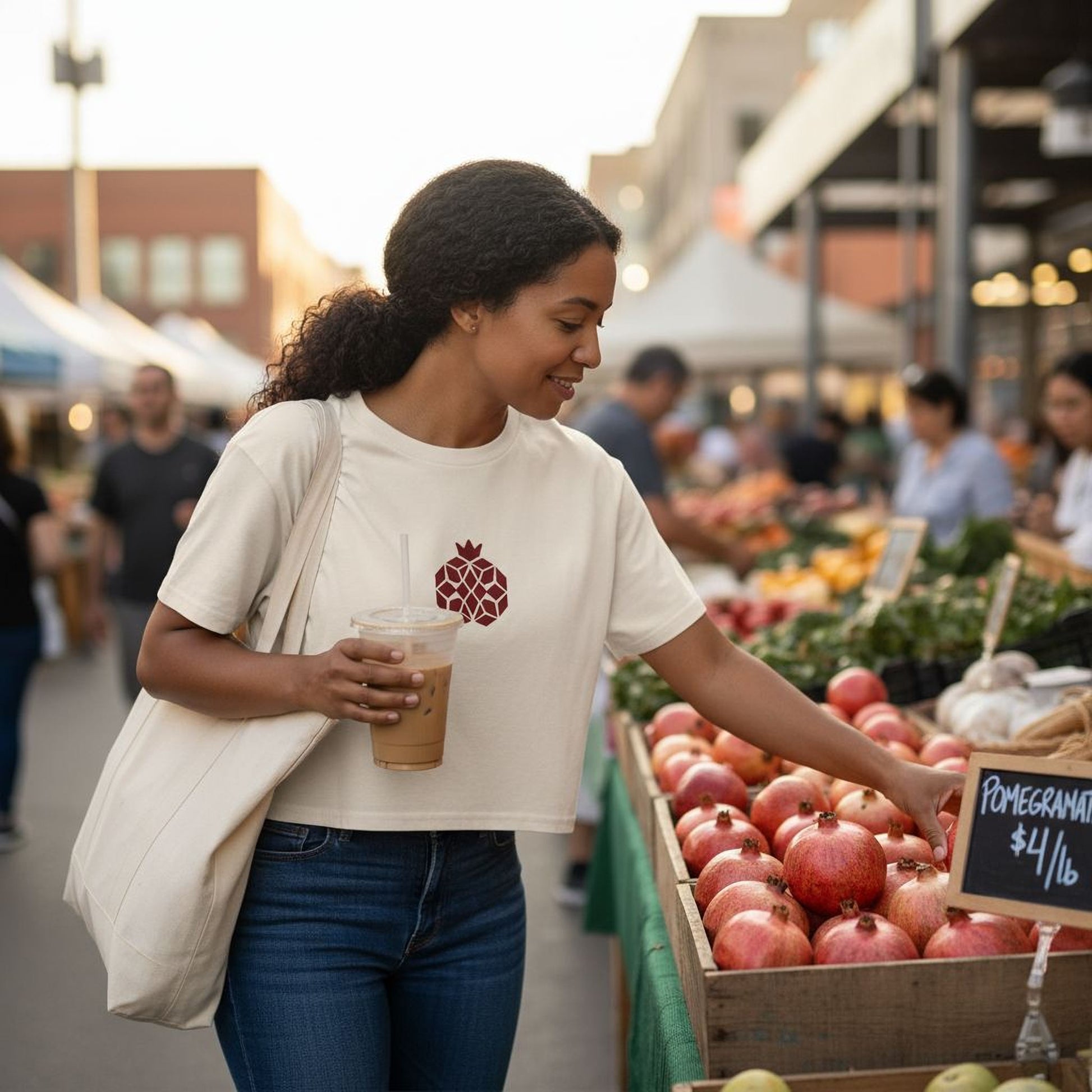A woman is shopping at an outdoor market, examining a selection of pomegranates for sale. She is holding a drink in her hand and appears to be focused on the produce.