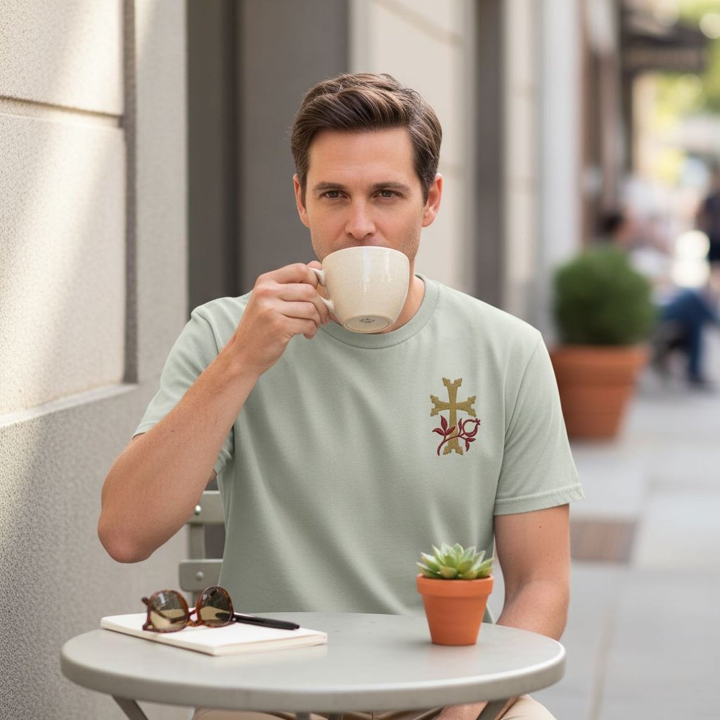 A man is sitting at a table outside, drinking from a cup and holding a pen in his hand. There is a potted plant and a small plant on the table, and a pair of sunglasses is also visible.