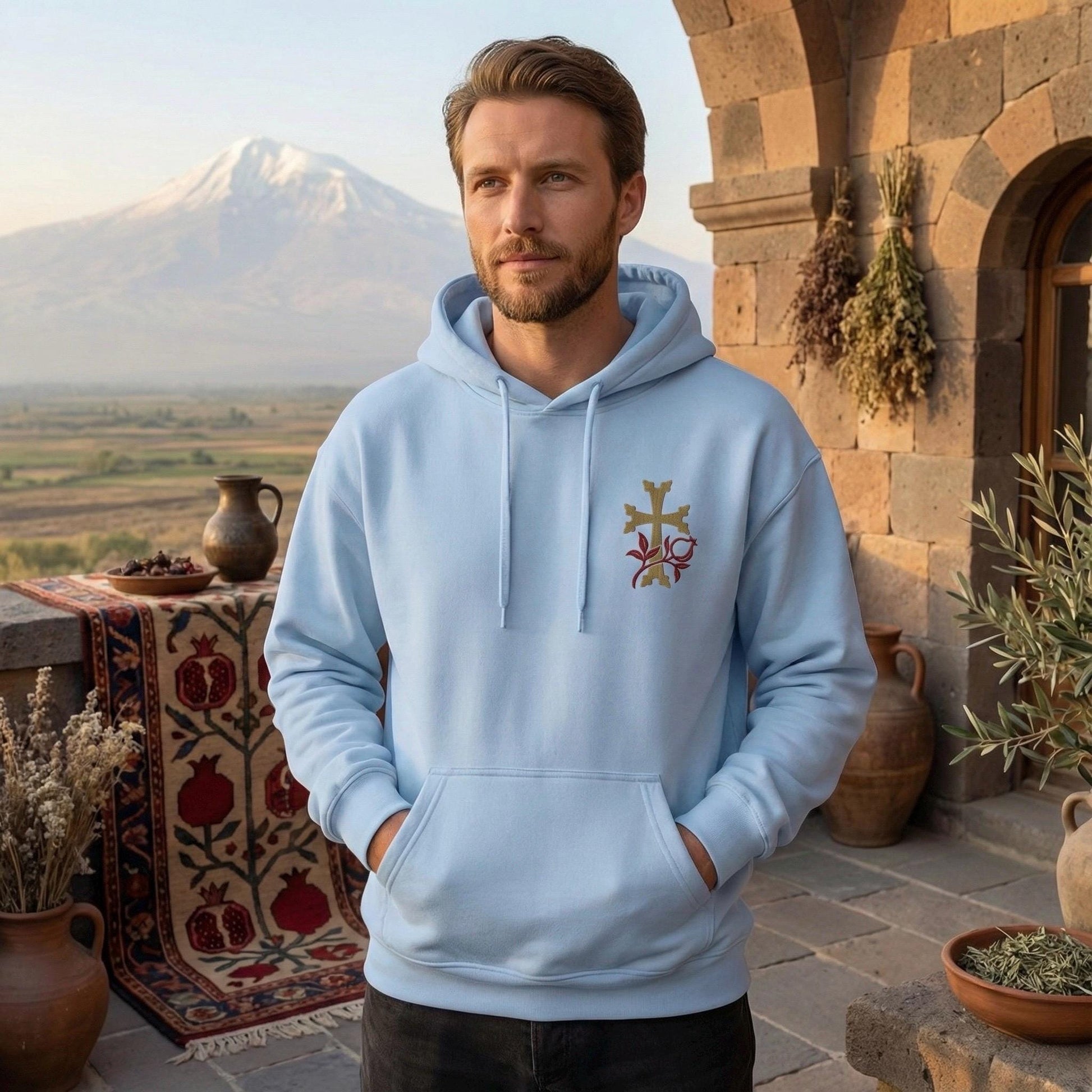 A man wearing a light blue hoodie with a cross design stands in front of a stone archway, with a mountain and a table with a pitcher and a bowl on it in the background.