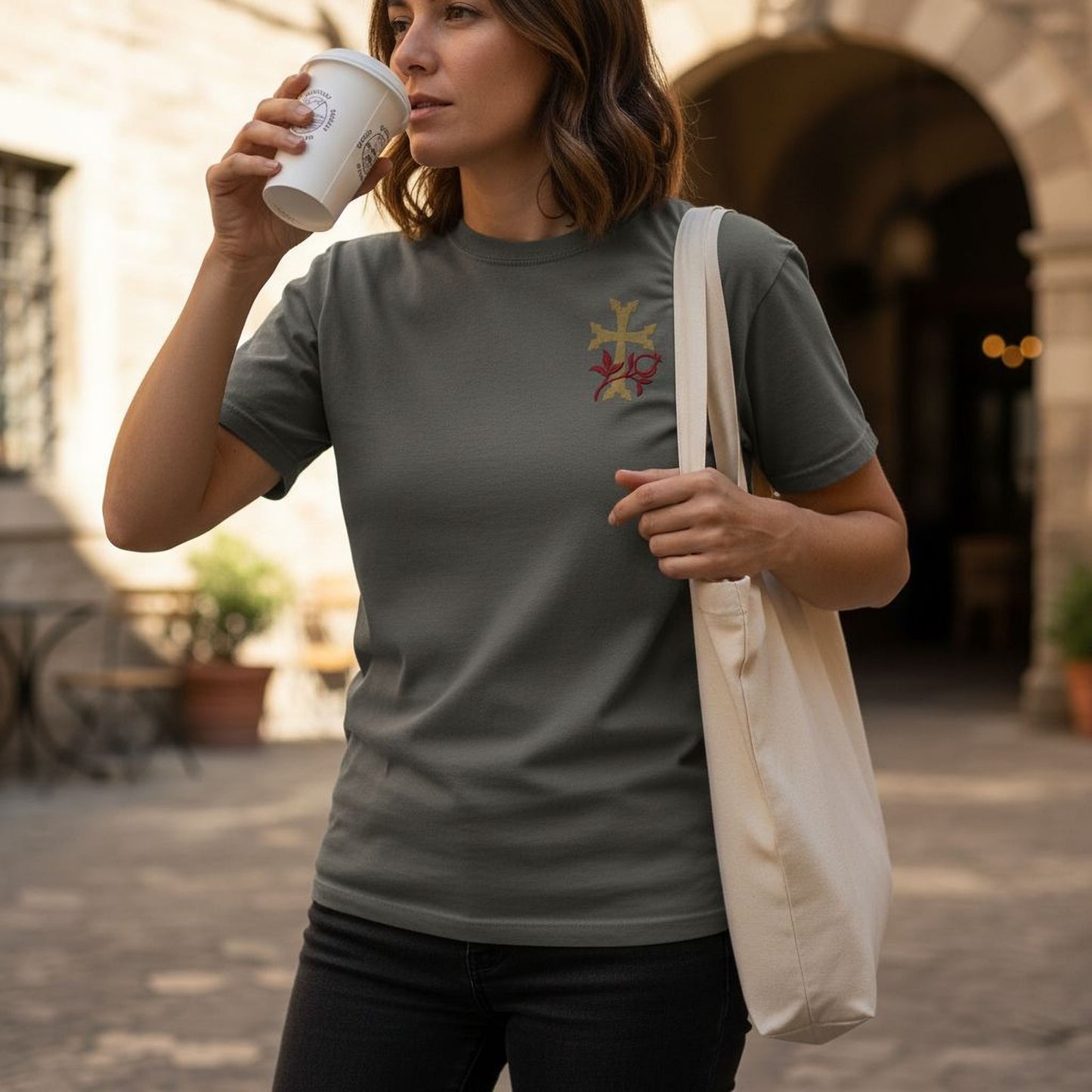 A woman wearing a gray t-shirt with a cross design and holding a white coffee cup is standing in front of a building.