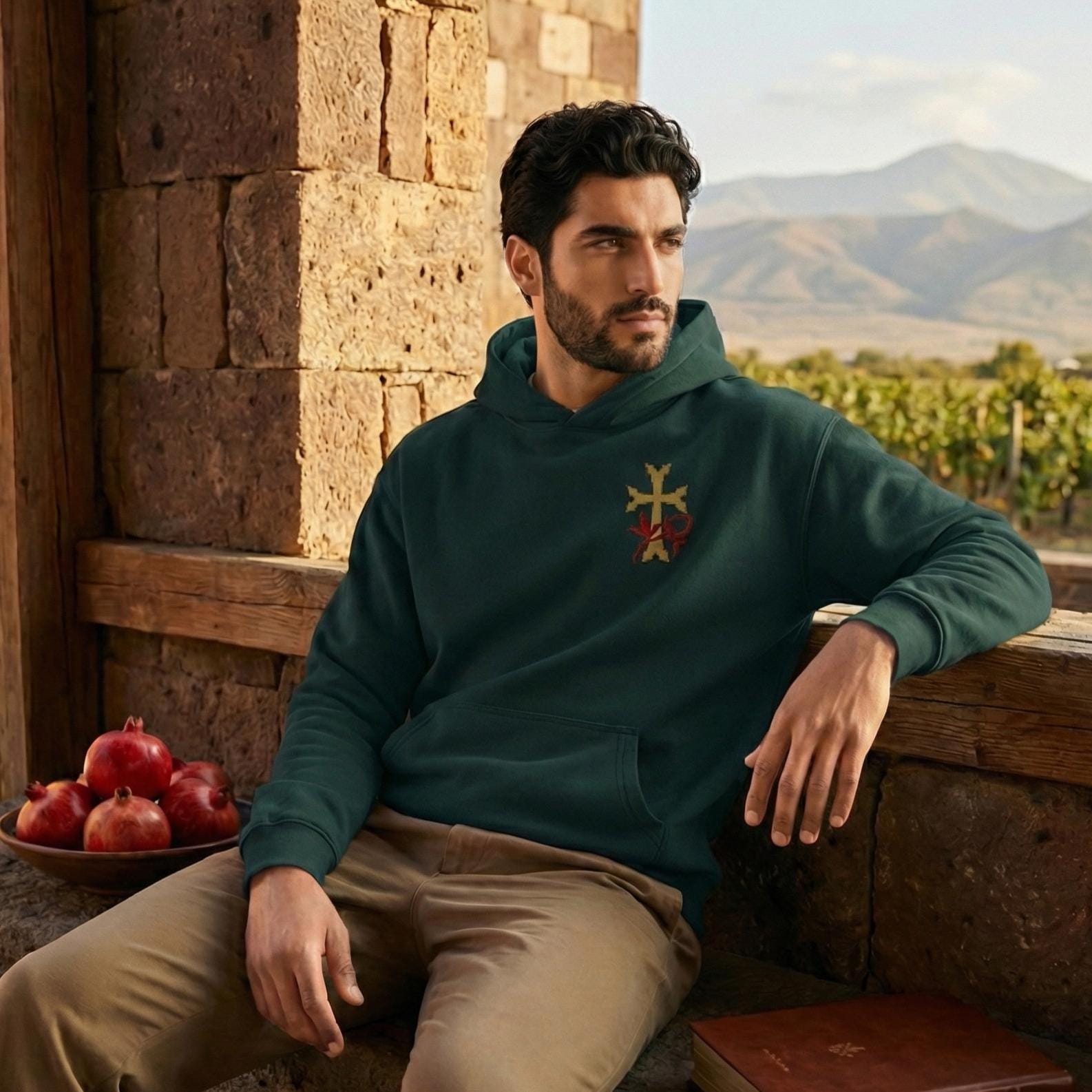 A man in a green hoodie with a cross on the front is sitting on a stone wall, leaning against it and looking off to the side. There are several pomegranates on a table in front of him.